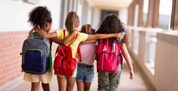 Rear view of multiracial elementary schoolgirls with backpacks and arm around walking in corridor. unaltered, childhood, together, education and back to school concept.
